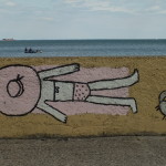 Graffiti na Bulwarze Nadmorskim w Gdyni. Kobieta Kobieta leży na brzuchu i się opala, głowę ma przykrytą kapeluszem przeciwsłonecznym. Obok rakwe